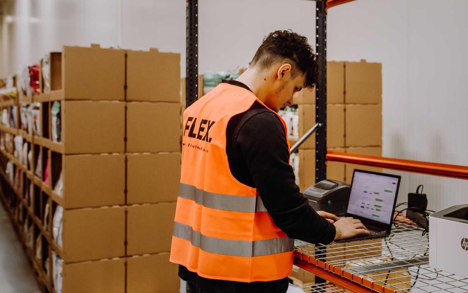A warehouse worker in an orange vest uses a laptop on a metal rack. Boxes line shelves in the background, creating a busy, organized atmosphere.