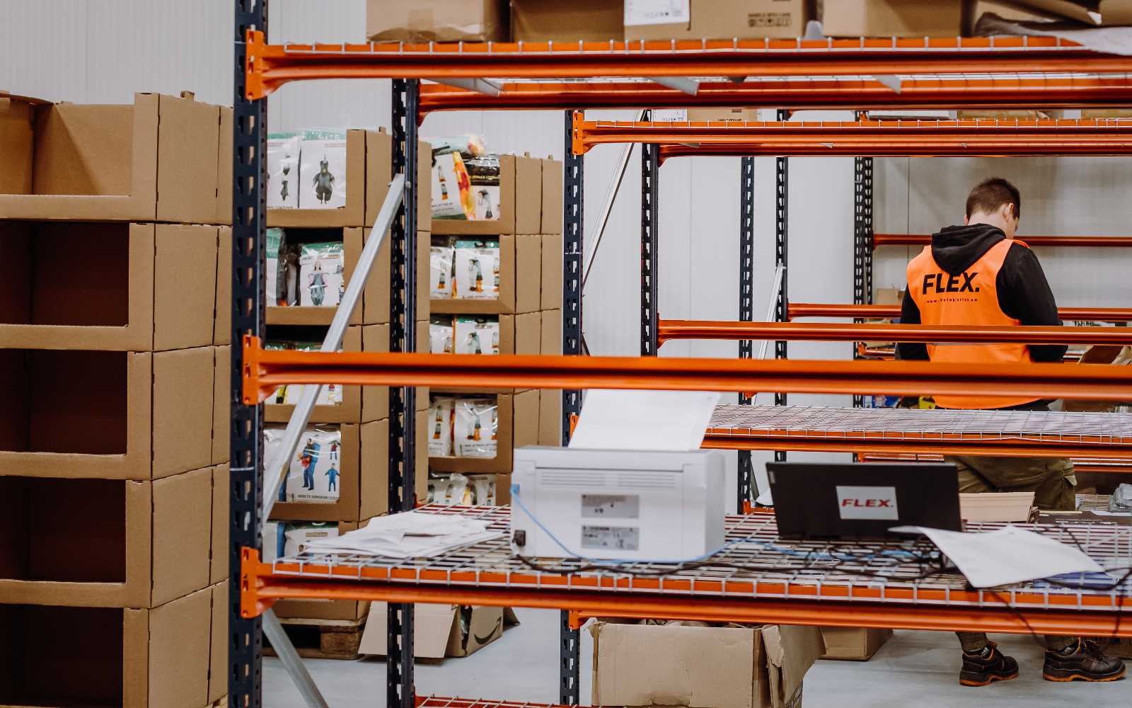 A person wearing an orange safety vest labeled "FLEX" handles a cardboard box in a warehouse. Shelves with more boxes are visible in the background. The tone is busy and industrial.