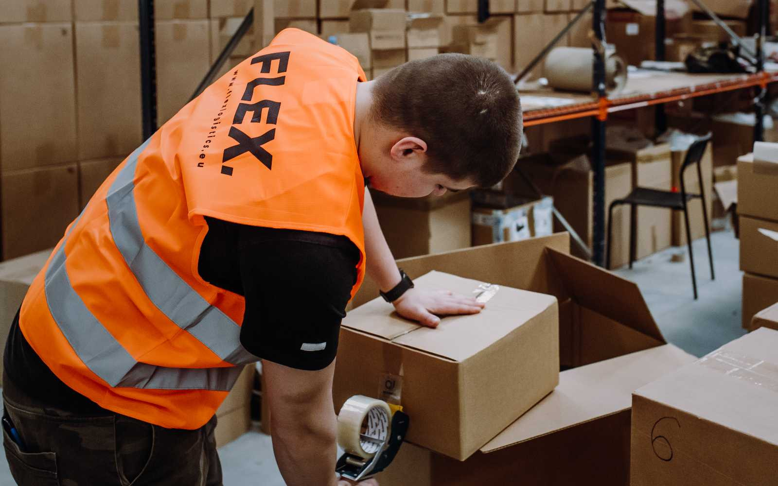 A person wearing an orange safety vest labeled "FLEX" handles a cardboard box in a warehouse. Shelves with more boxes are visible in the background. The tone is busy and industrial.