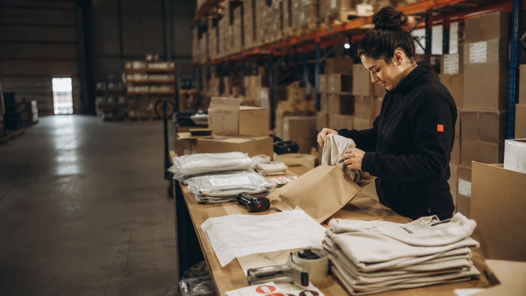 Warehouse operator packing folded apparel into mailers at a fulfillment station with shelving and cartons in the background.