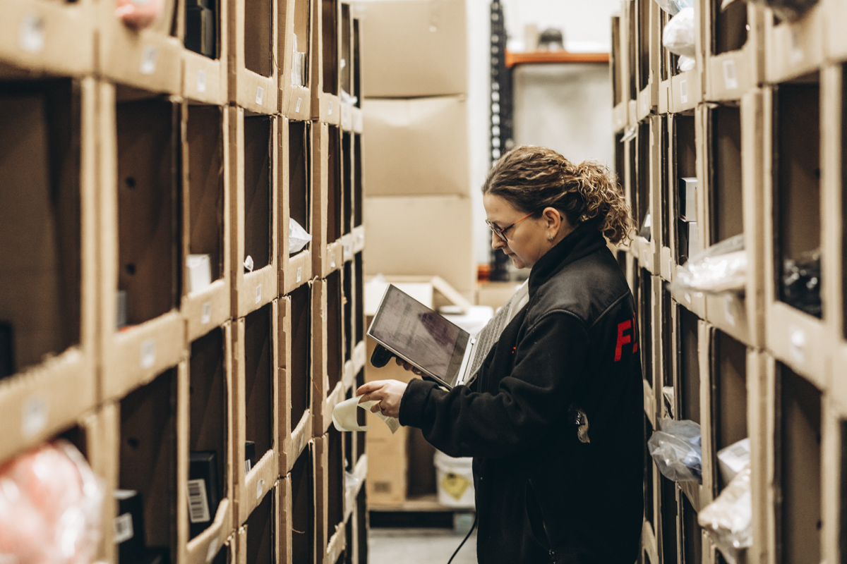 FLEX. warehouse worker reviewing inventory information on a tablet while checking storage locations in a warehouse shelving aisle.